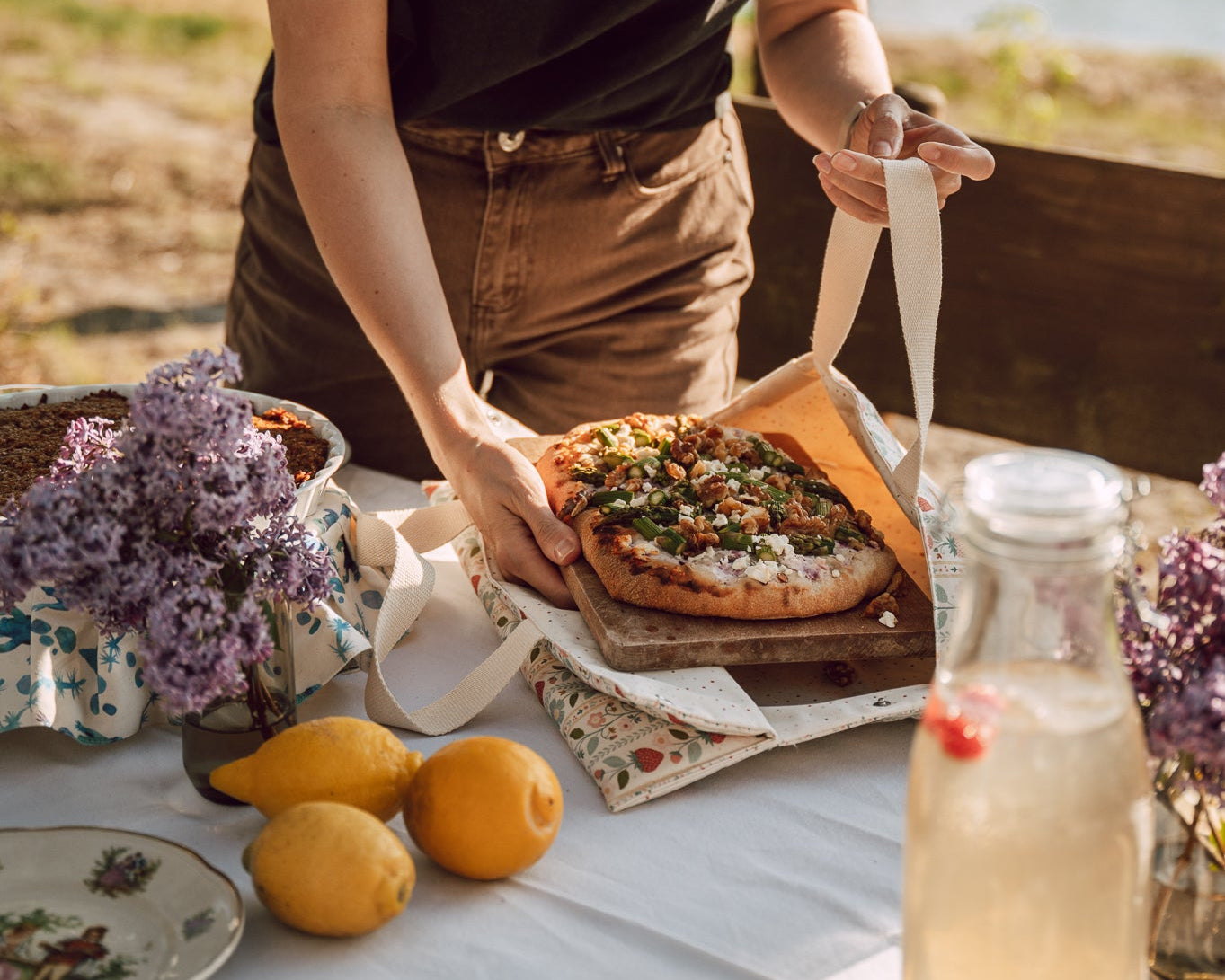 A woman in a black t-shirt and brown pants is serving a pizza from a wooden box on a white tablecloth set outdoors near a lake, with lemons, lilacs, and a glass of lemonade in the foreground.