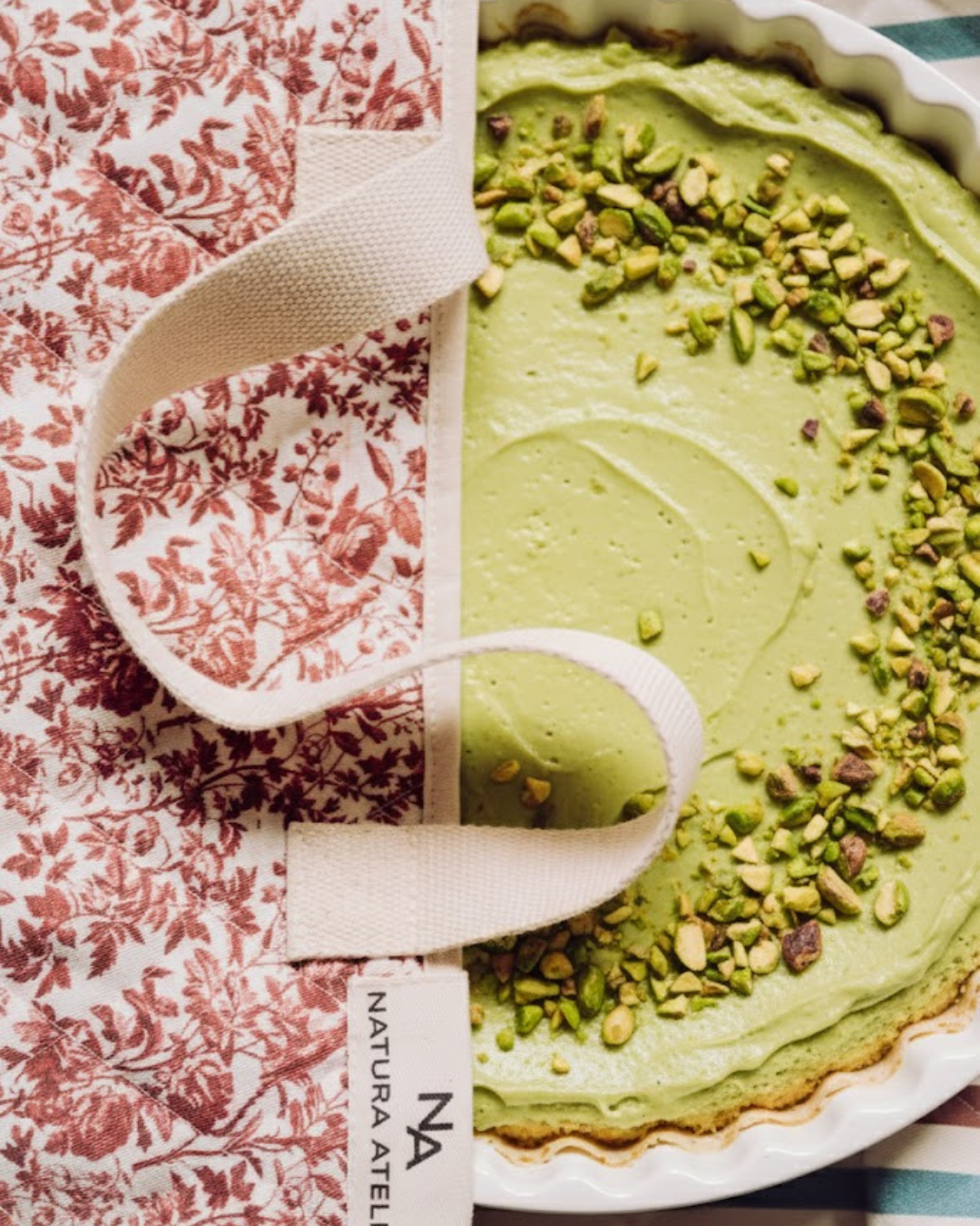 Green dessert with pistachio nuts in a white dish in Toile de Jouy cake carrier bag , with 'Natura Atelier' branding.