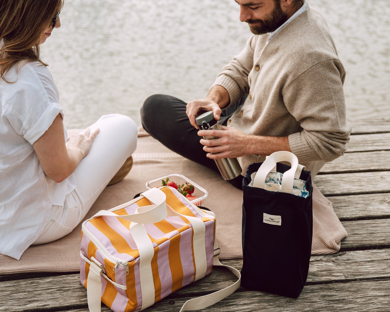 A man and a woman sit on a wooden dock by the water, with picnic bags in front of them. One bag is orange and white striped, and the other is black.