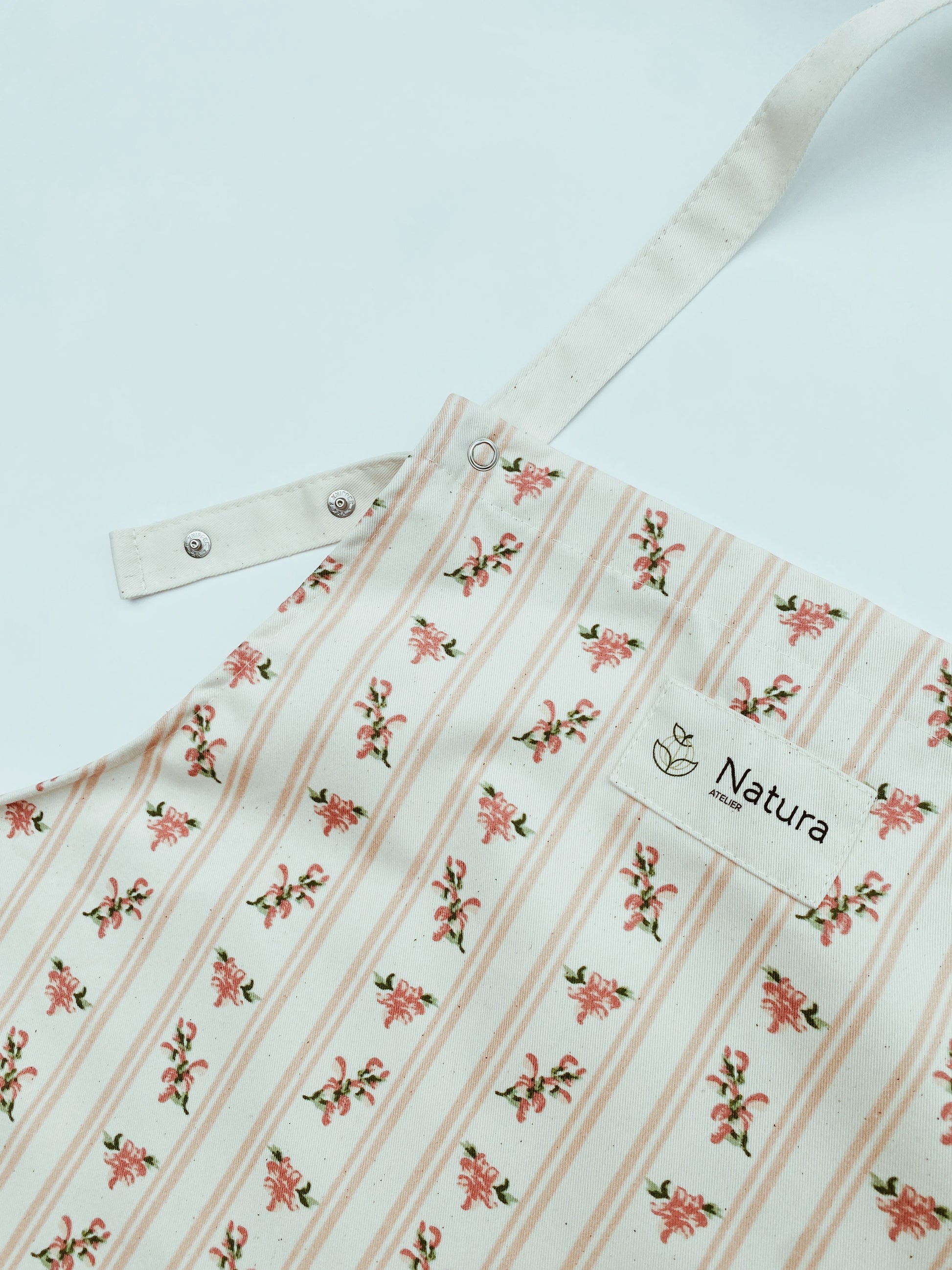 Floral-patterned apron with 'Natura Atelier' branding on a light blue background